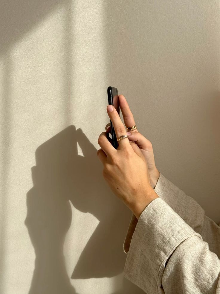 A hand holding a phone against a sunlit linen-coloured wall, casting a long shadow.